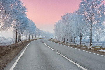 A frosty road curves through a winter landscape under a vibrant pink and purple sunset sky, with snow-dusted trees on both sides.