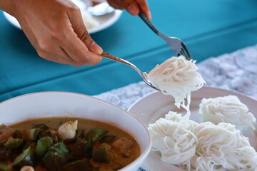 Women use spoons and forks to scoop rice noodles from the tray onto a plate. The rice noodles are arranged into bite-sized balls for easy eating. Popularly eaten with green curry is placed on the side