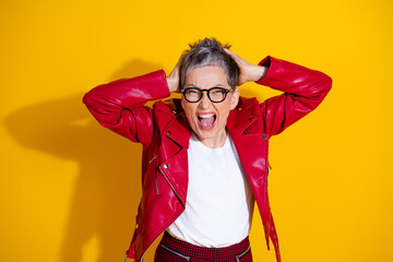 Cheerful senior woman in a stylish red leather jacket expressing happiness with hands on head...