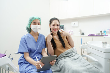 Fototapeta premium Female patient smiling and pointing at cheeks while sitting next to Asian dental assistant in blue uniform, both posing happily together in examination room after consultation session in dental clinic
