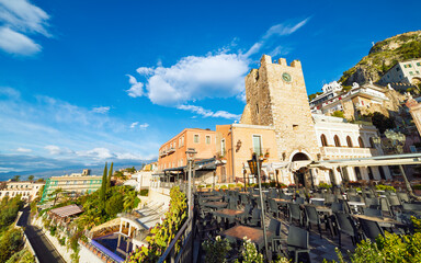 Stone clock tower and empty outdoor terrace in Taormina old town, Sicily. Bright morning sunlight, traditional Mediterranean architecture and scenic hillside view create classic Taormina atmosphere