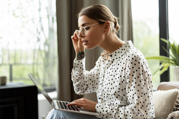 Focused freelancer woman adjusting glasses while working on laptop at home