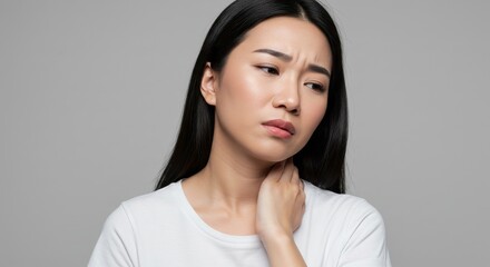 Portrait of an Asian woman touching her neck with a pained expression, suggesting sore throat, swollen lymph nodes, or neck pain. Neutral gray background. Concept related to healthcare, illness, and p