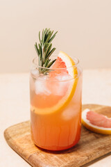 Grapefruit cocktail with rosemary garnish in a glass, served on a wooden cutting board. Vibrant, refreshing drink perfect for summer menu or cocktail recipes. Greyhound, lemonade, paloma, mocktail.