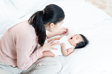 Asian mother gently looking at newborn baby yawning while lying on bed. Baby dressed in white onesie with arms relaxed. Moment conveys tiredness, bonding, daily care, and peaceful affection.