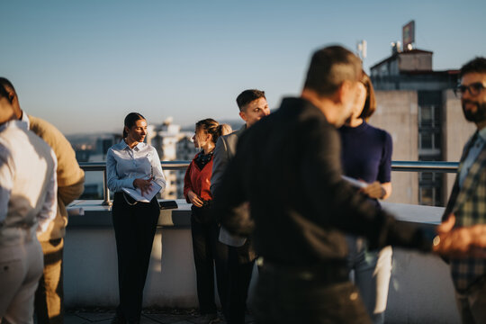 A group of multiracial business people engage in a creative brainstorming session on a high-rise rooftop at sunset, fostering collaboration and innovation in a relaxed outdoor setting.