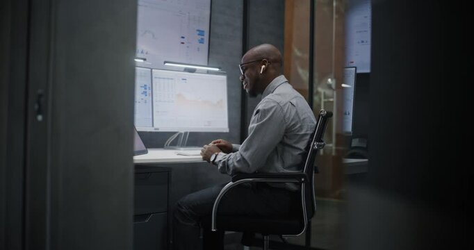 Stock Exchange Trader in Wireless Headphones Having Call, Monitoring Real-Time Stocks, Exchange Market Charts on Multi-Monitor Desk Setup. African American Investment Agent Working in Office at Night.
