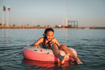 Smiling girl relaxing on a pink inflatable donut in the sea at sunset. Summer vacation fun, joyful...