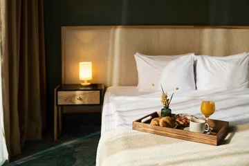 Fotobehang Hotel chique Cozy hotel room showcased with breakfast tray on bed. Tray filled with coffee, juice, pastries, and fruit, with warm lamp providing ambient lighting next to bed  © pressmaster