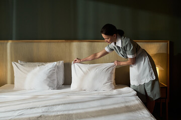 Hotel housekeeper making bed in a clean, modern room, arranging pillows on neatly made bed, wearing...