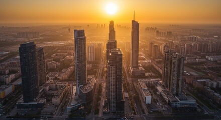 Fototapeta premium Aerial view of a cityscape at sunset featuring modern skyscrapers silhouetted against a warm orange sky. The sun's rays create a dramatic lighting effect over the urban landscape.