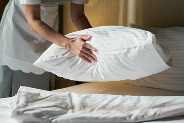 Housekeeper arranging fresh white linens and fluffing pillow in a guest room. Focus on neatness and attention to detail while preparing comfortable sleeping environment