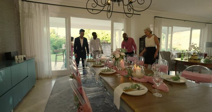 Following host gesturing, Diverse friends entering dining room, taking seats at pink-sashed chairs