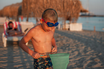 Young boy wearing swim goggles playing on sandy beach with a green net. Warm golden hour light and...