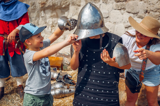 Children and adults trying on medieval armor outdoors, laughing and helping each other, participating in a fun historical reenactment or educational event.