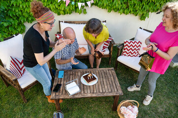 Overhead view of joyful senior friends clapping and smiling around birthday cake during outdoor family gathering with gifts and decorations