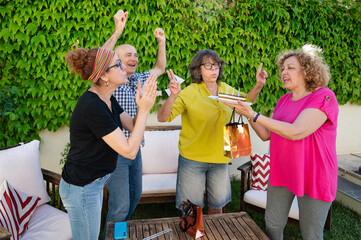 Middle aged woman blowing candles on birthday cake held by friend while joyful group cheers and claps in vibrant outdoor garden celebration