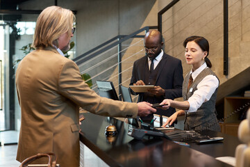 Businessman in beige suit interacting with hotel reception staff while checking in at front desk. Receptionist scanning card while another staff member taking notes on clipboard