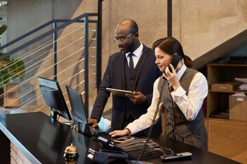 Professional team engaged in work at a contemporary office, using multiple devices and discussing project details at front desk. Dynamic setting with a blend of technology and human interaction