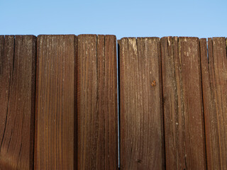 Close-up of a vertical wooden garden fence against a clear blue sky. Weathered planks show natural texture, simple structure, and rustic outdoor design
