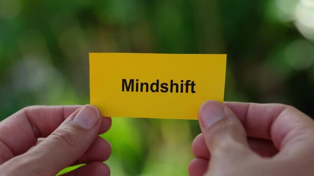 A woman holding a yellow paper note with the word Mindshift written on it in her hands against a foliage background. Close up.