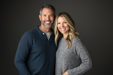 A happy couple, mid-aged, smiling at the camera, with a gray background wearing casual sweaters.