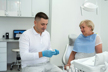 Obraz premium dentist holds an implant and consults an elderly woman with the installation of a tooth crown, dental clinic