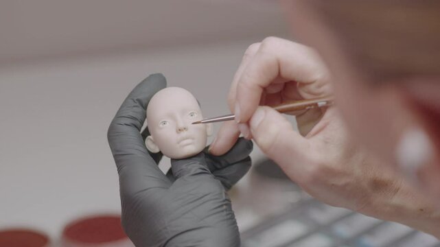 Artist carefully adds fine details to silicone doll head using thin brush, highlighting precision and craftsmanship in miniature figure creation
