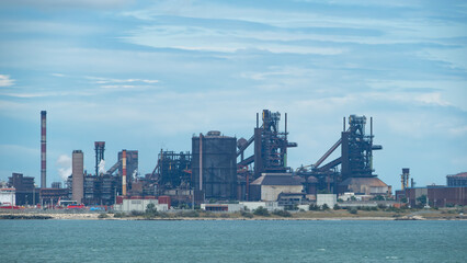 .Industrial steel plant with smokestacks and complex structures, seen across water under a cloudy sky