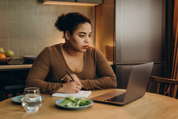 African American woman makes notes listening to webinar via laptop at table in kitchen, eating...