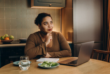 Satisfied African American woman writes healthy recipes from laptop at round table in kitchen