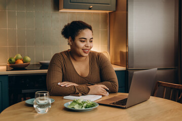 Smiling woman makes notes listening to webinar via laptop at table in kitchen