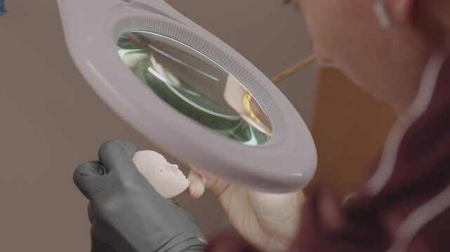 Closeup of artist painting silicone doll head through magnifying glass while wearing gloves, showing careful process of fine detailing in miniature creation