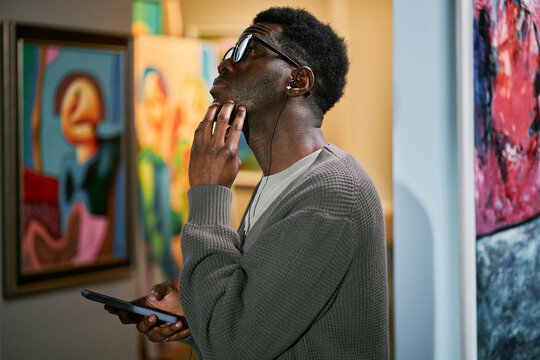 Man with headphones looking at painting in art gallery while holding phone. Engaging in thoughtful contemplation while surrounded by vibrant artwork in gallery