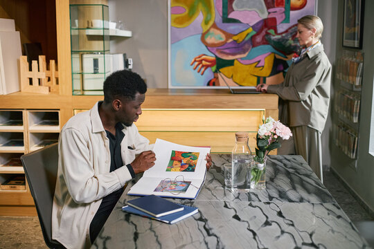 Two artists in a studio discussing colorful painting, one seated at table with a sketchbook, other standing near artwork on wall. Creative tools and flowers are on table