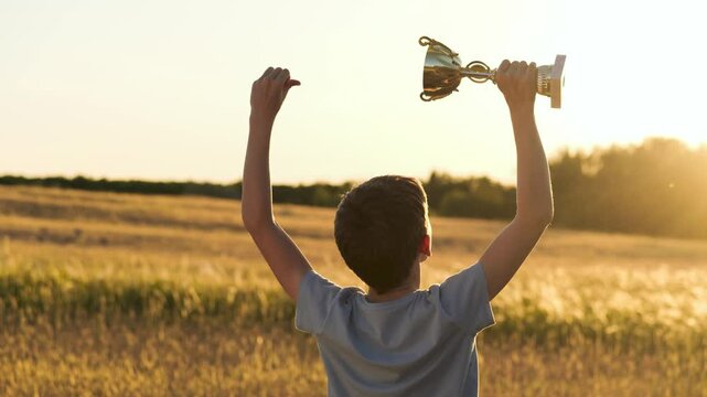 boy raises golden cup high into sky. silhouette boy with his arms raised and golden cup. silhouette of winner triumphant cup in rays of sunset. boy winner celebrates trophy