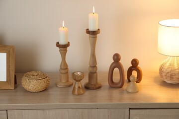 Stylish candlesticks with burning candles and other decor on cabinet near beige wall indoors