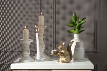 Elegant candlesticks with burning candles, branches in vase and other decor on dresser near grey...