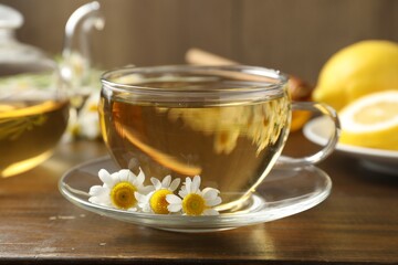 Delicious chamomile tea in glass cup, fresh flowers and lemon on wooden table, closeup