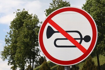 Close-up of a no honking traffic sign with a horn crossed out in red. The round sign stands against a background of green trees and a partly cloudy sky.