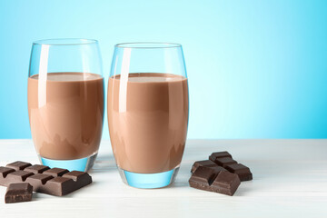 Tasty chocolate milk in glasses and pieces on white wooden table against light blue background