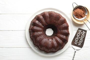 Tasty chocolate bundt cake, cocoa powder and server on white wooden table, flat lay. Space for text