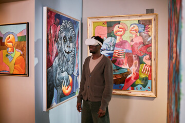 African American man wearing virtual reality headset standing between colorful paintings in...
