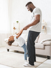 Black dad teaching little naughty daughter dancing at home, girl staying on father feet and having...