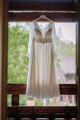 Elegant white wedding dress with lace detail hanging outdoors on a wooden balcony with soft natural light and greenery in the background..