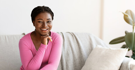 Positive young african american woman posing on sofa at home, resting on comfortable couch in cozy...