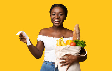 Cheerful young african american woman holding supermarket bill and bag with groceries after successful shopping, planning food budget, bought organic fruits and vegetables, posing on yellow background
