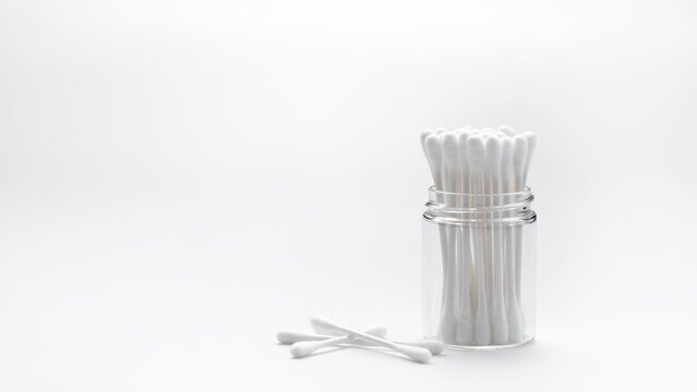 A clear glass jar filled with cotton swabs is placed on a white surface, with a few loose cotton swabs lying in front of it.