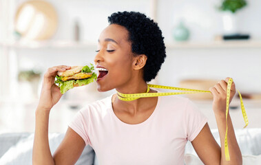 African American woman wanting to eat cheeseburger, pulling herself away by tape measure at home,...