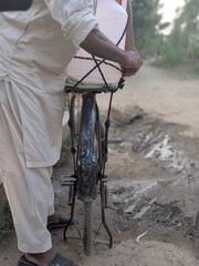 person standing on dirt road holding bicycle, wet weather, overcast sky, rural landscape, detailed,...
